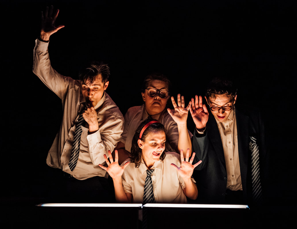 Image: A group of three are pretending to be pressed up against glass, a woman crouching below them. There is a low-light below them illuminating their faces, and there is no other light onstage. All are wearing what looks like school uniforms and have exaggerated, hungry facial expressions.